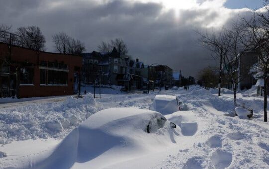 In the corner, a sliver of blue sky is seen next to a large, dark cloud. Underneath, cars parked along a commercial street, appear to be only humps of snow.