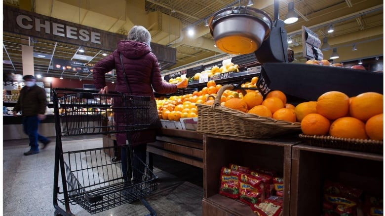 A woman in a coat shops for oranges in a supermarket in St. John's. Behind her, a cheese display is shown.
