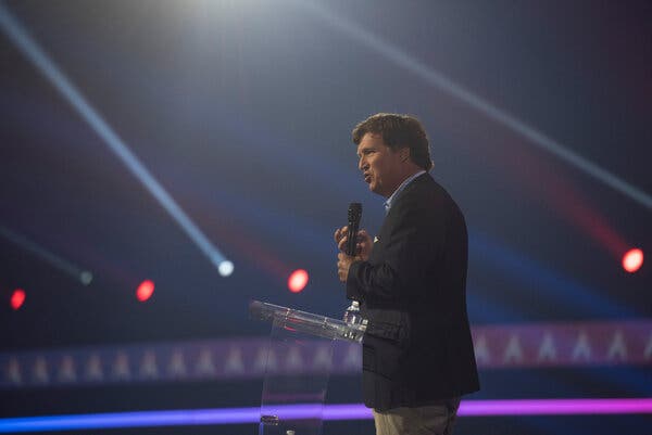 Tucker Carlson stands on a stage with a microphone in hand and neon lights in the background.