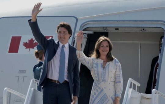 A man and a woman stand on a stairway outside the entrance to an airplane and wave.