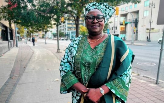 A woman in a green traditional Nigerian outfit stands on a sidewalk and smiles